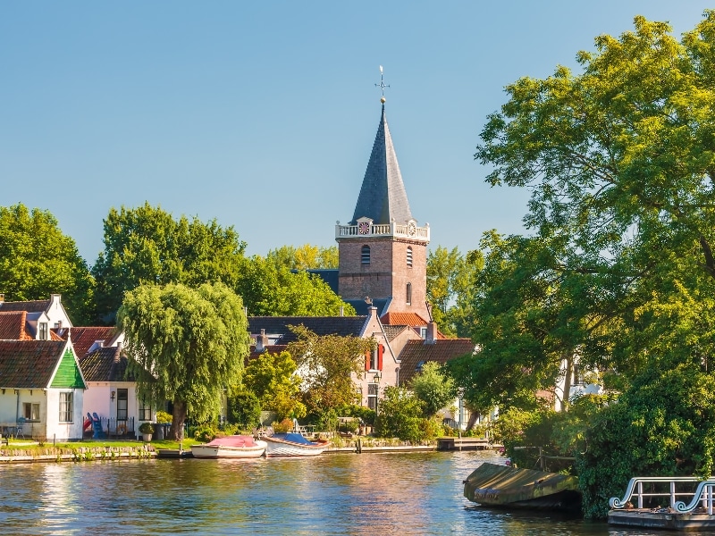 small square church tower topped with a spire in front of a canal lined with houses, boats and trees