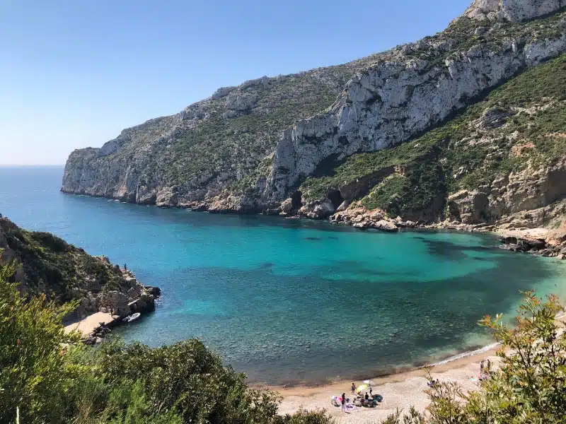 sandy beach with vegetation covered cliffs to either side and a small group of poeple looking out to the clear turquoise sea