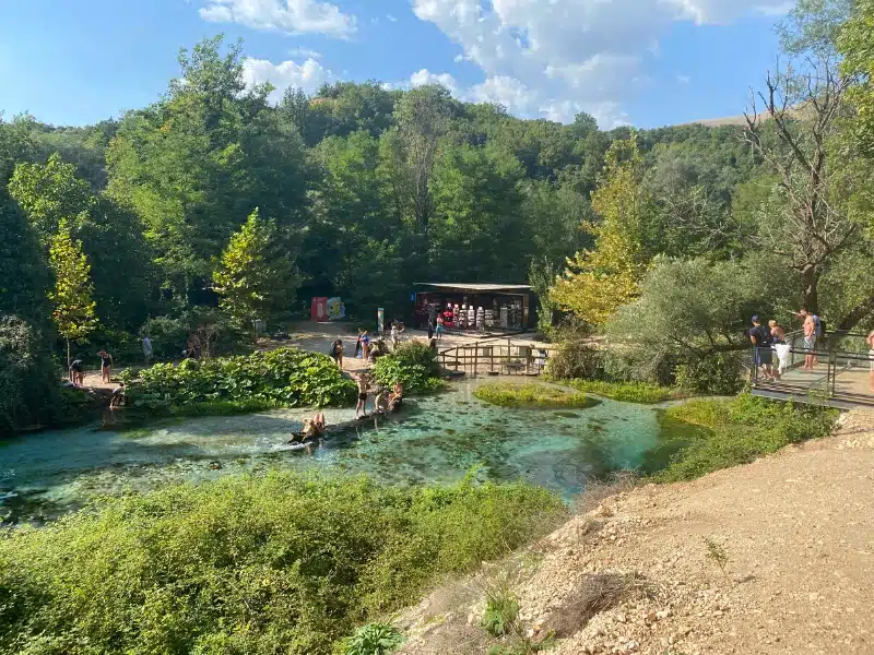 a natural spring pool surrouned by greenery with a small building in the distance and people sitting and standing around the water