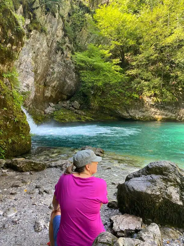 Woman in a pink tshirt and grey hat sitting on a rock in front of a natural pool with a waterfall.