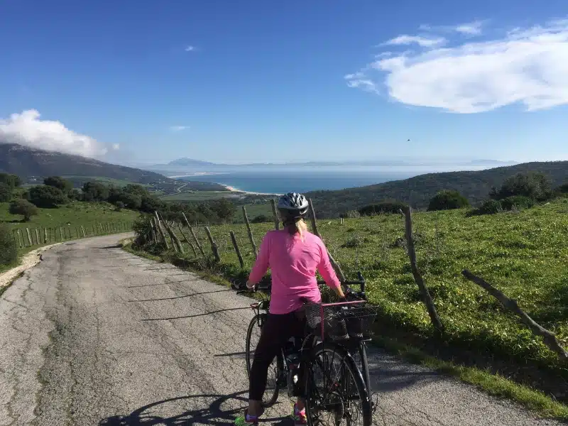 Woman astride a bike wearing a silver bike helmet with a distant view of sea, beach and woods