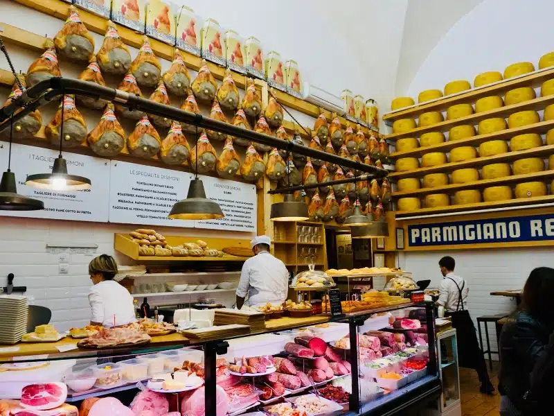 shop with a deli counter and staff serving, with the white tiled walls behind lined with wheels of parmesan cheese and whole Parma hams