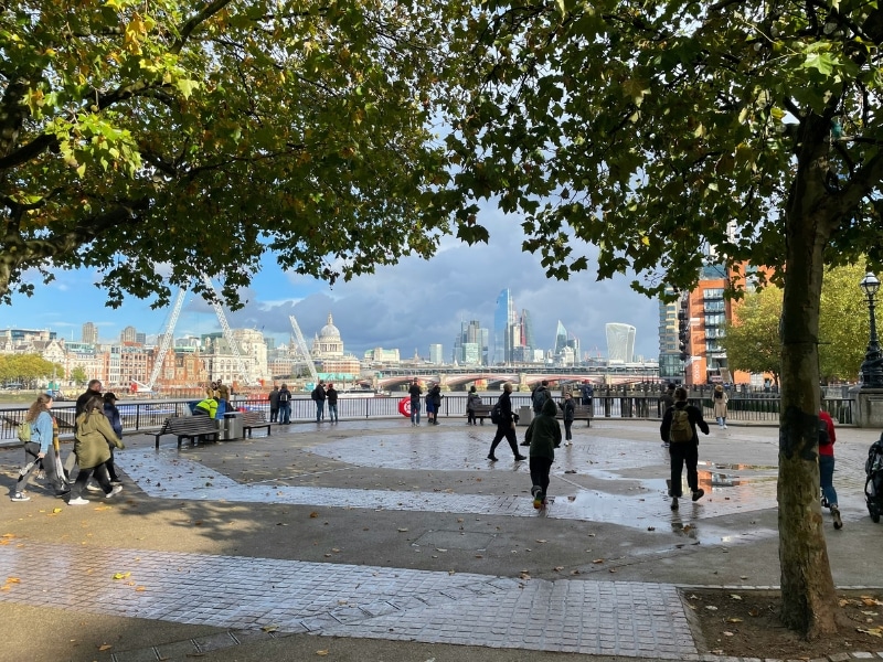 square full of people under leafy trees with a view of a city over a river