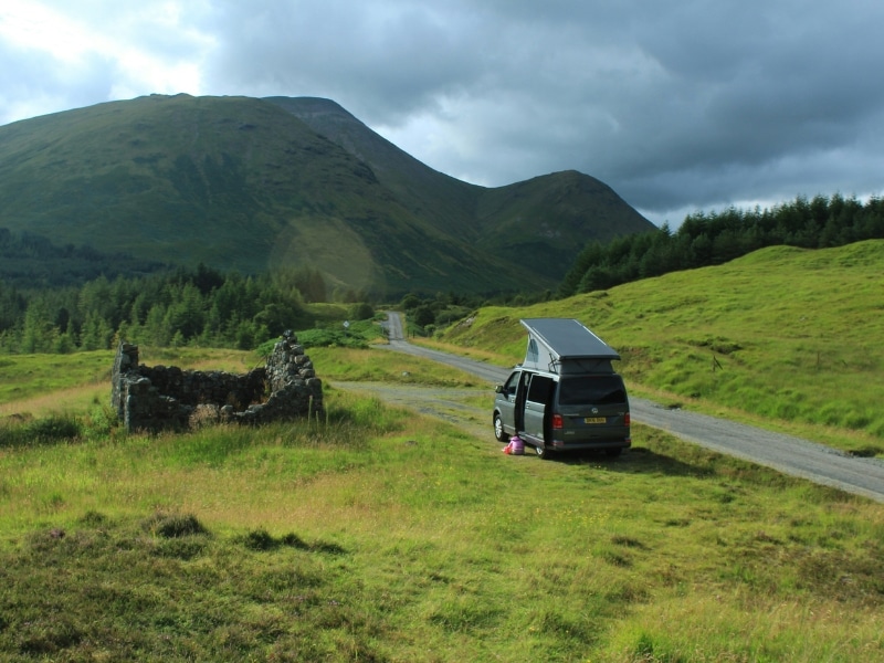 a green campervan parked in a grassy meadow beside a derelict stone building and road