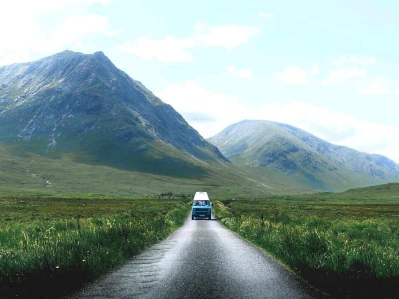 blue and white campervan driving along a road lined with tall grasses, with mountains in the background