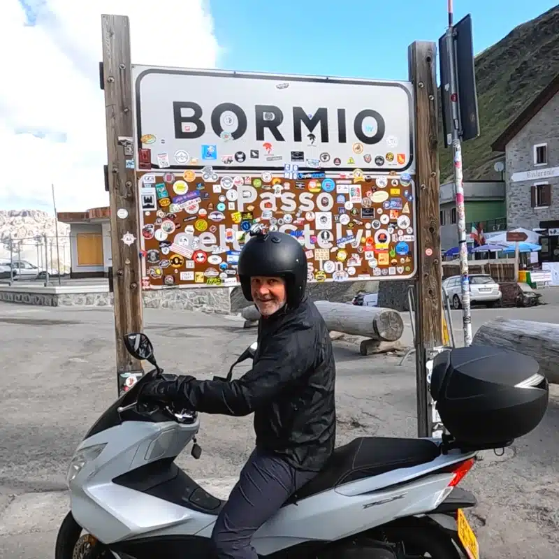 man on a moped wearing a black helmet by a 'Bormio' sign covered in stickers