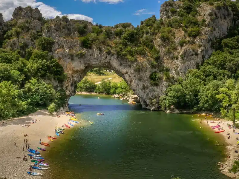 Natural rocky arch over a shallow river flanked by beaches with people and river craft on them