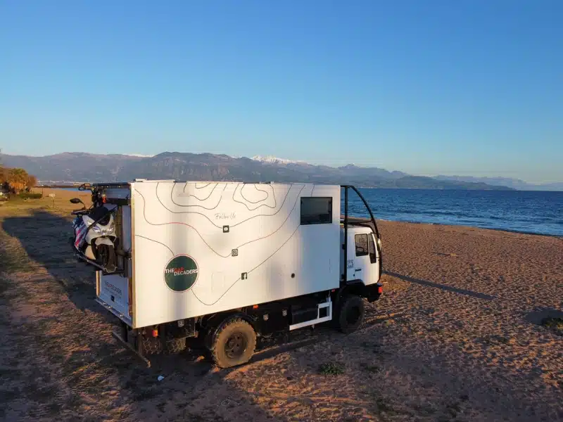 white overland truck parked on a beach with the sea and mountains in the background