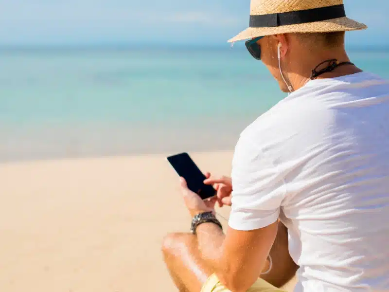 man wearing a straw hat and white t-shirt sitting on a beach using a mobile phone