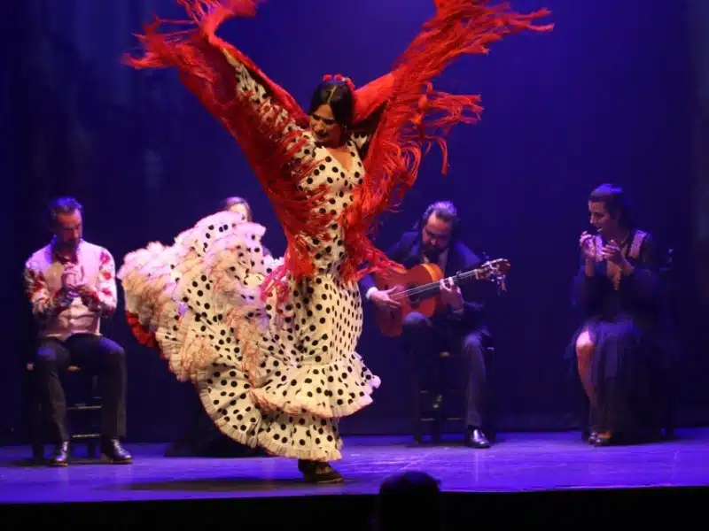 woman in black spooted dress and red shawl dancing on a stage surrounded by guitarists