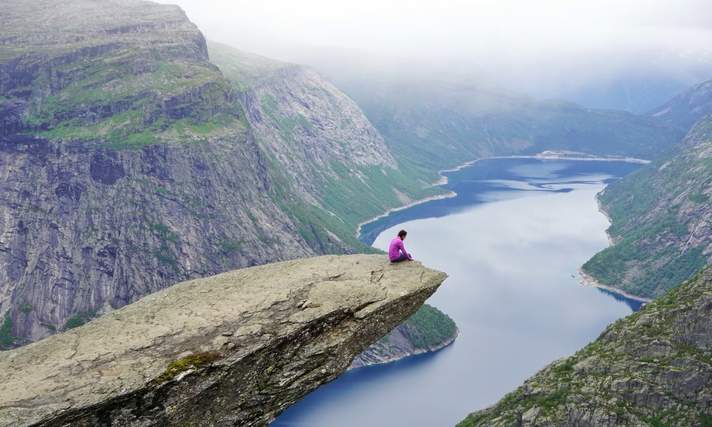 young woman in a pink top sitting on the edge of a high rock overlooking a lake