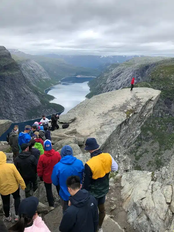 line of people in colorful clothes waiting to walk onto a large rock jutting out over a lake
