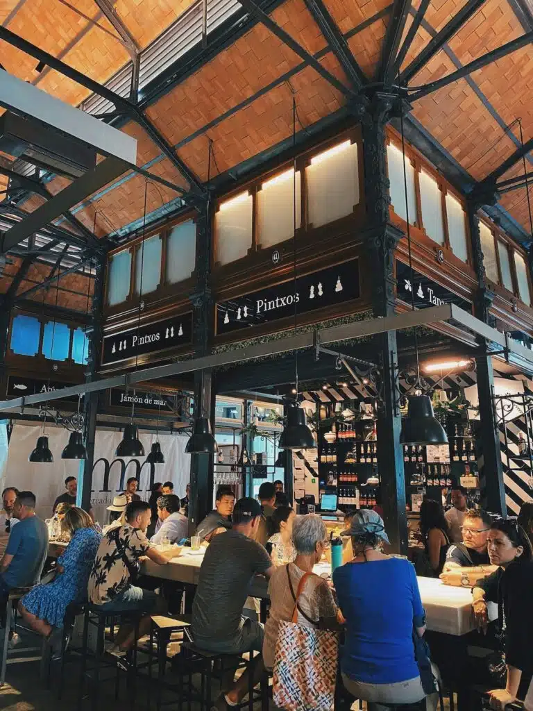 inside a large food market in Spain with people sitting on stools at a pinxo bar