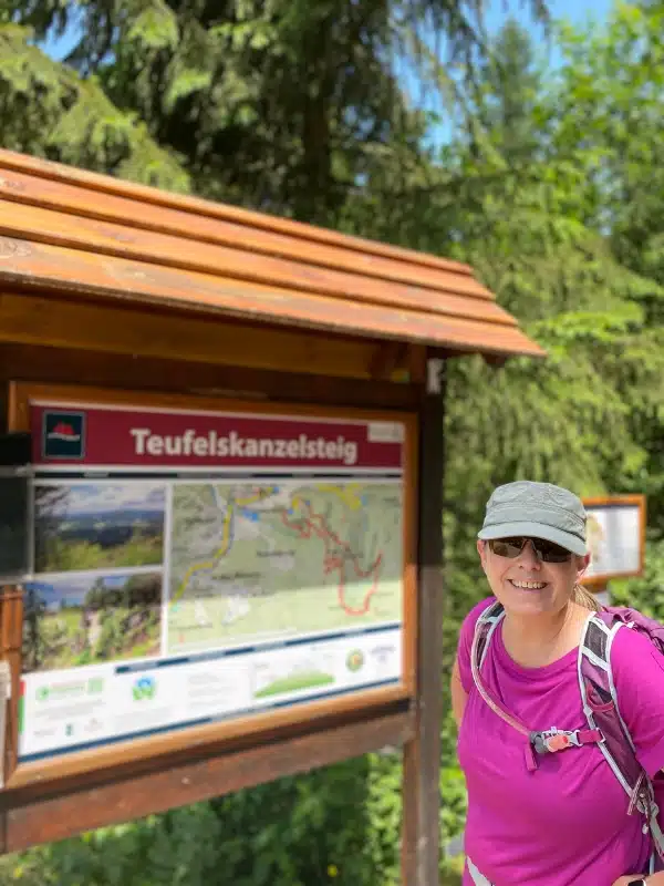 woman in a pink t-shirt and grey cap standing by a hiking map on a wooden sign covered with a small roof