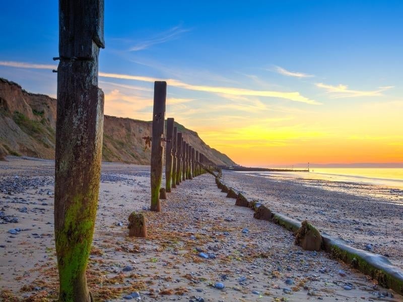 shingle and sand beach at sunset backed by small cliffs with wooden poles along the beach