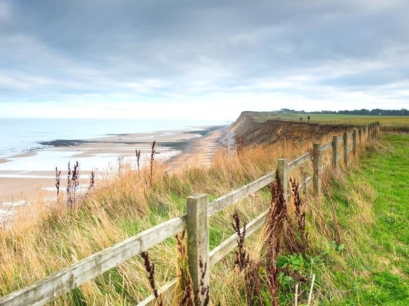 large beach backed by a sandy cliff taken from a high position in a grassy field with a wooden fence