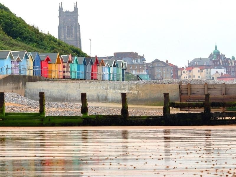 A row of colourful wooden beach huts facing a sandy beach with a town and church tower in the backgroun