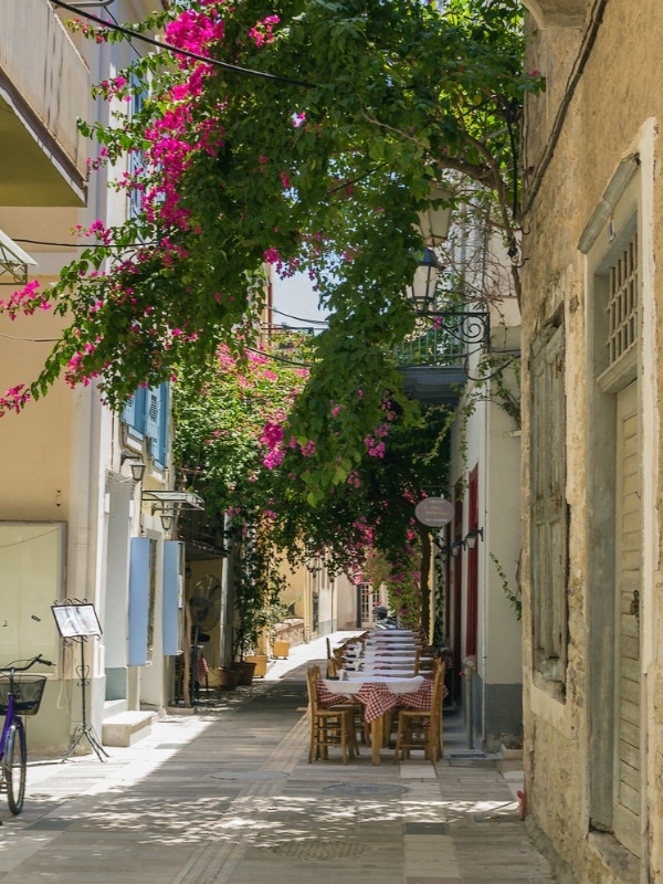 stone buildings covered in bougainvillea lining a narrow street with cobbles and red check tablecloth covered tables in the distance