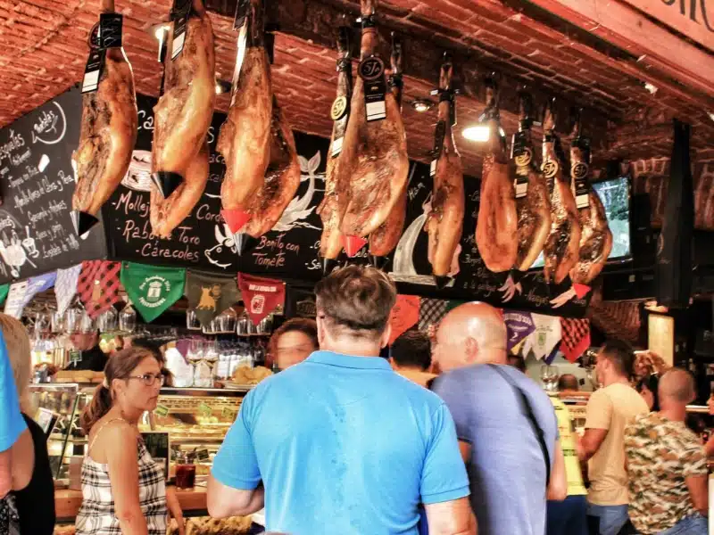 people in a bar surrounded by jamon hanging from the ceiling and other Spanish tapas