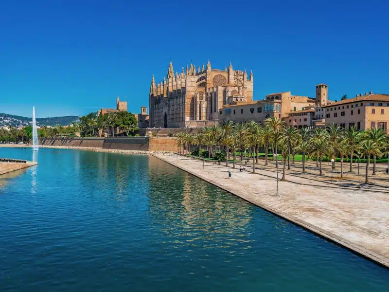 artificial lake with a fountain surrounded by stone pavements and palm trees with a large stone cathedral in the background