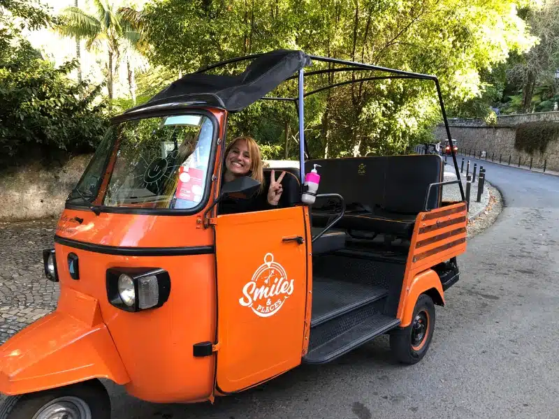 woman in an orange tuk-tuk with trees and stone walls in the background