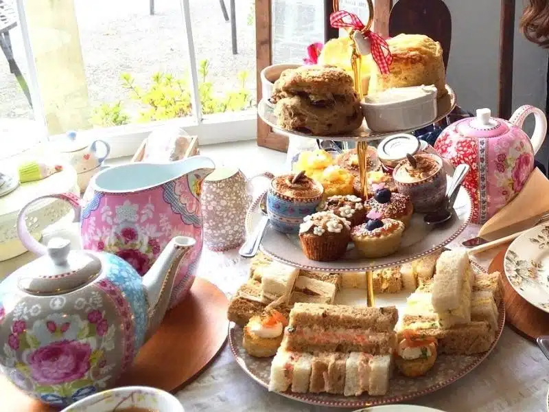 Table laid for afternoon tea with cake stand full of cake and sandwiches and colourful crockery and teapot