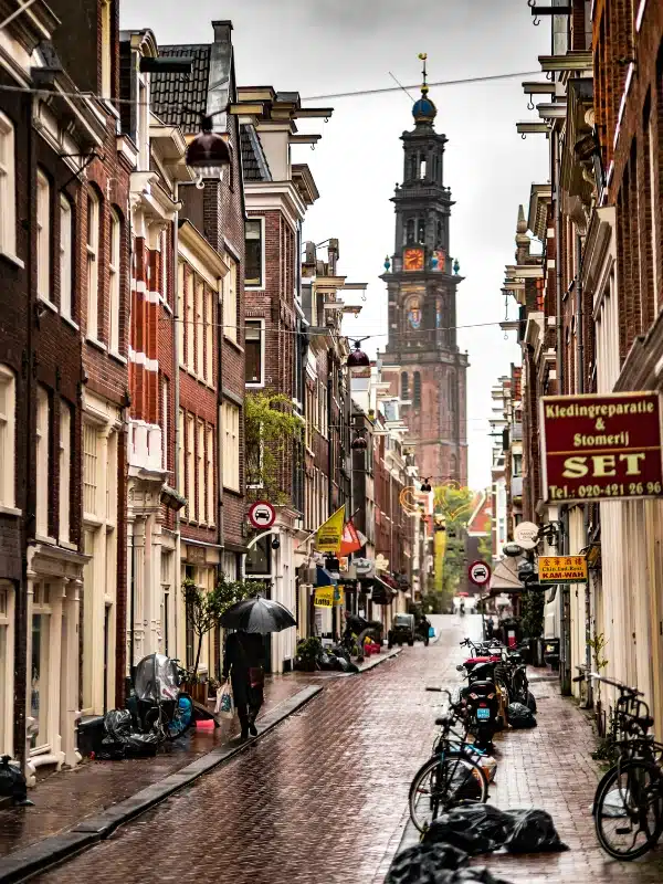 narrow street lined with bicyles and aman walking with an umberalla. There is an elegent church tower at the end of the street.