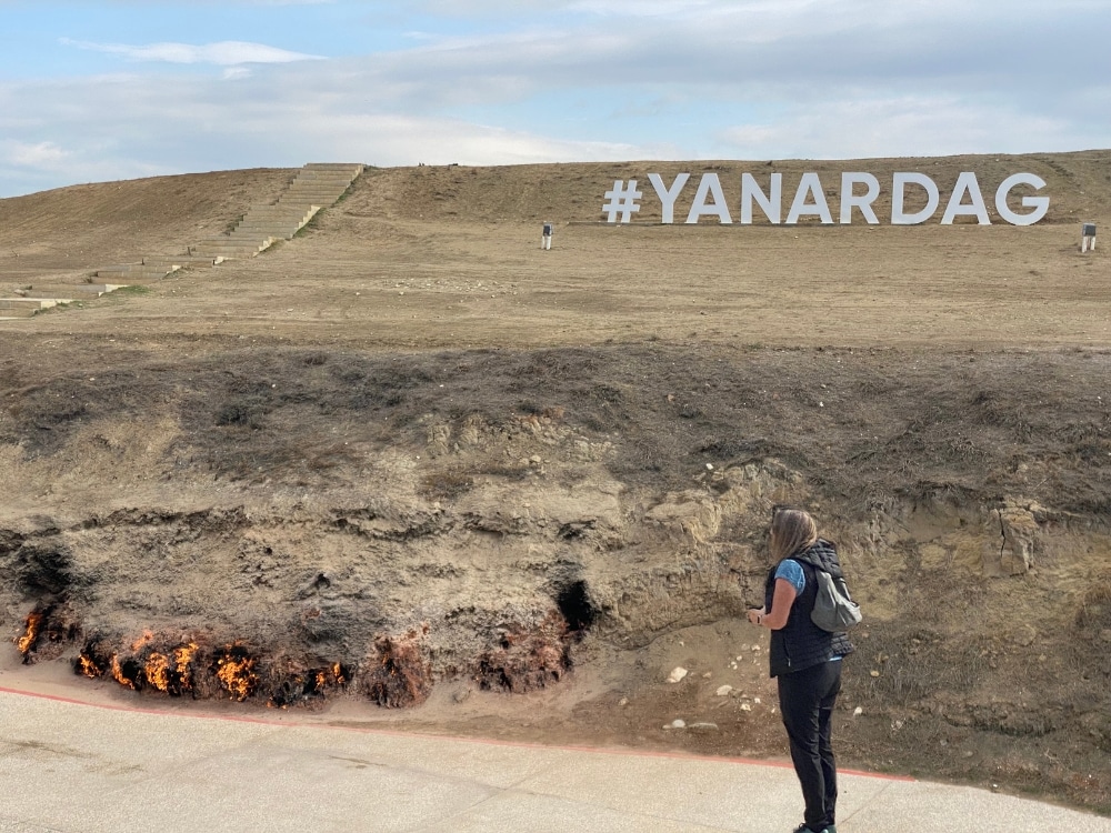 woman standing by a natural gas outlet in a hillside with a white #YANARDAG sign on the hill