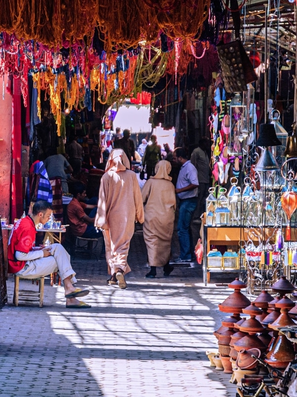 two robed men walking through a souk with brightly colored wool hanging from the roof