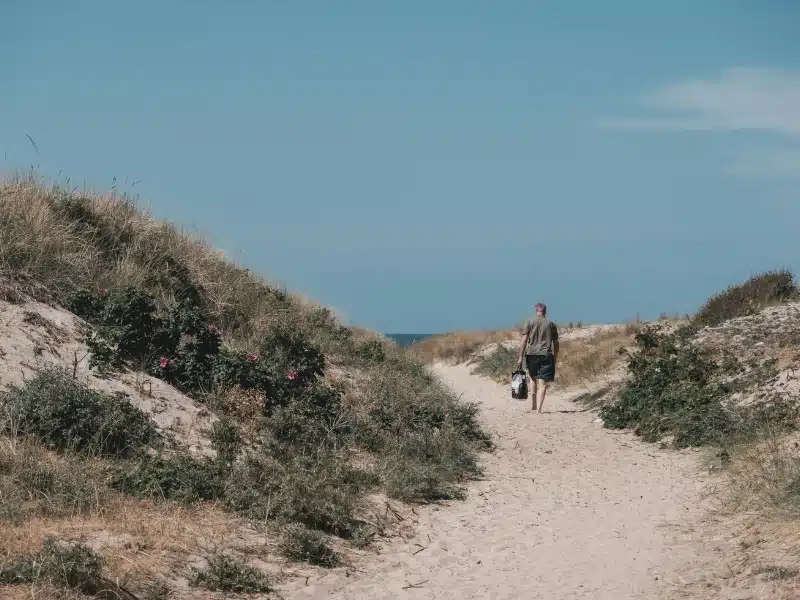 man walking along a sandy path with a glimpde of the sea through grassy dunes
