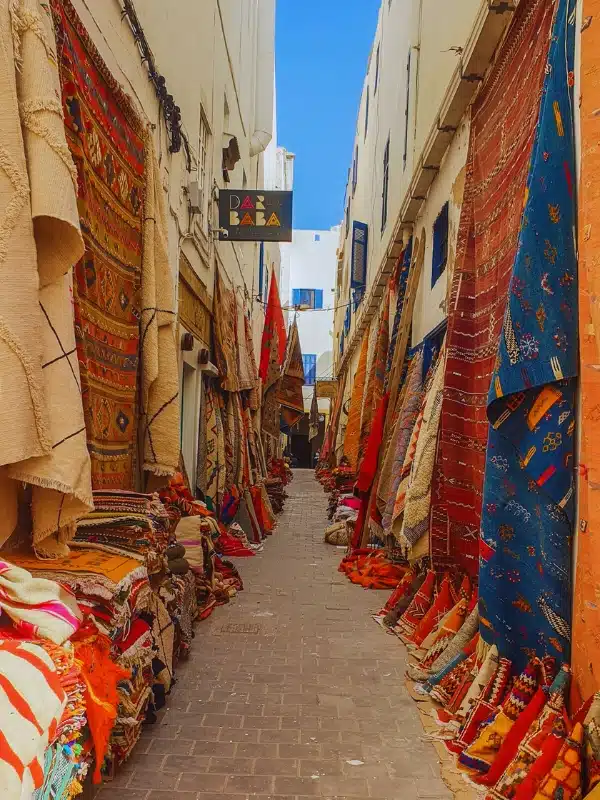 colourful handmade rugs hanging along high walls lining a narrow pagges with a white and blue building at the end