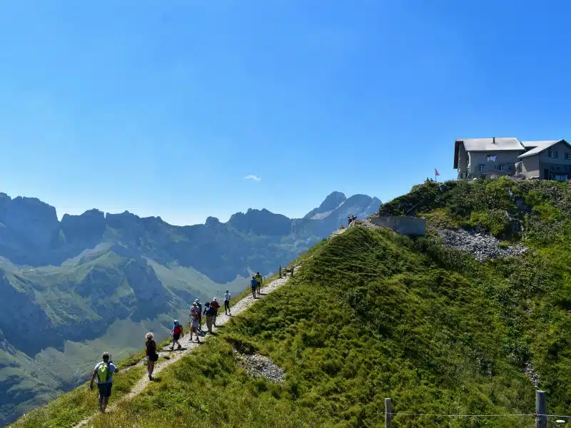 people walking along a mountain path to a refuge with a blue sky and other large mountains in the background