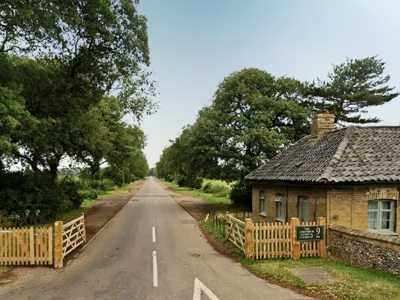 long road lined with grass and trees and a wooden fench and brick cottage at the start of the road