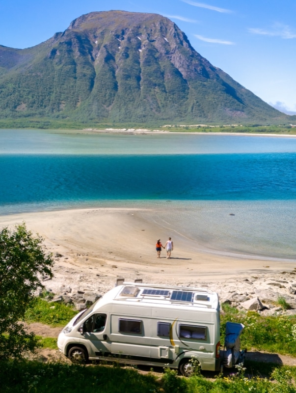 couple walking on a sandy beach with vibrant blue sea with a campervan parked in the foreground