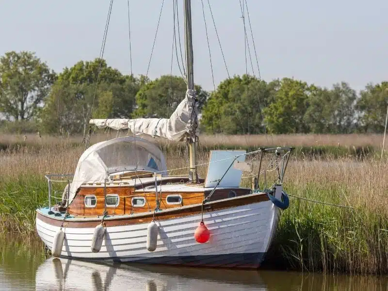 small sailing boat with sails stowed, moored in a river by a field on grass