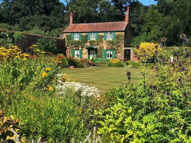 small cottage with green window shutters behind a lawn and flower beds