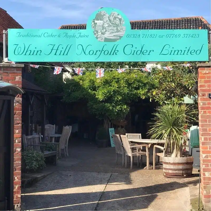 large turquoise sign hanging over brick pillars to a courtyard with 'Whin Hill Norfolk Cider Limited' printed on it