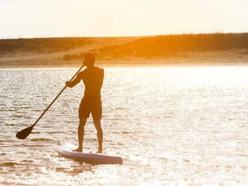 man on a stand up paddle board at sunrise in calm waters with the shore in the distance