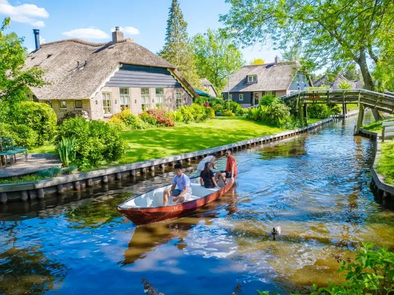 people in a boat on a canal in a Dutch village, with houses and a bridge in the background