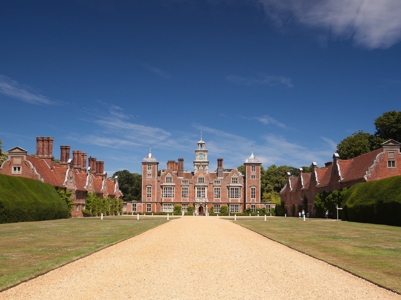 Jacobean building with turrets and many windows with a gravel path leading to it lined with grassed lawn