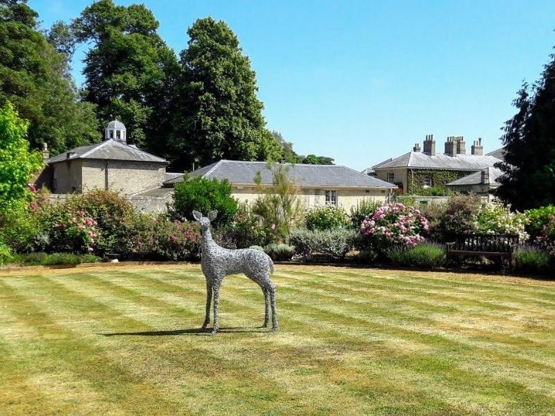 Sculpture of a deer on a lawn with buildings and flowers in the background