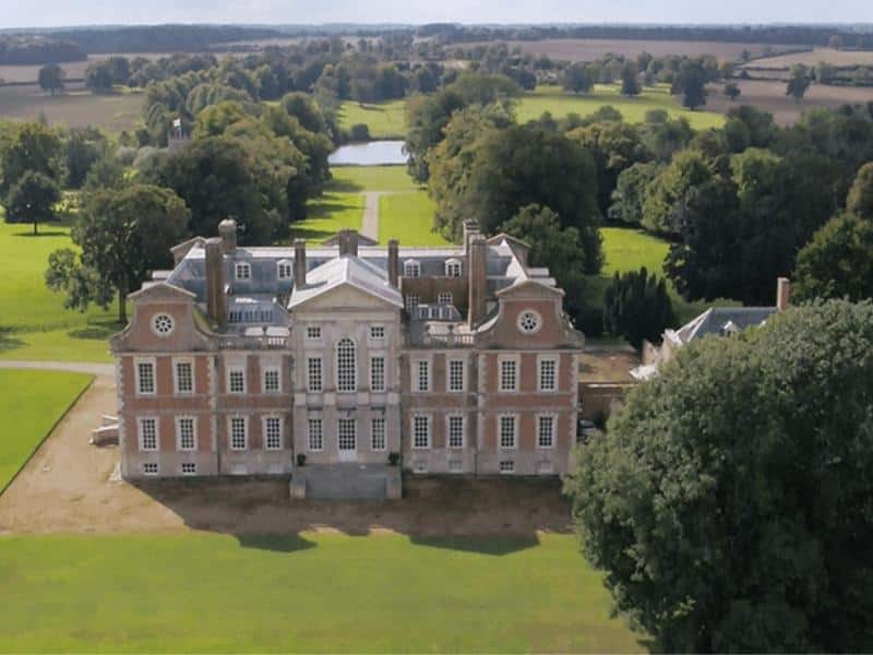 Large historic building with many windows set in a large informal garden of grass, trees and alake