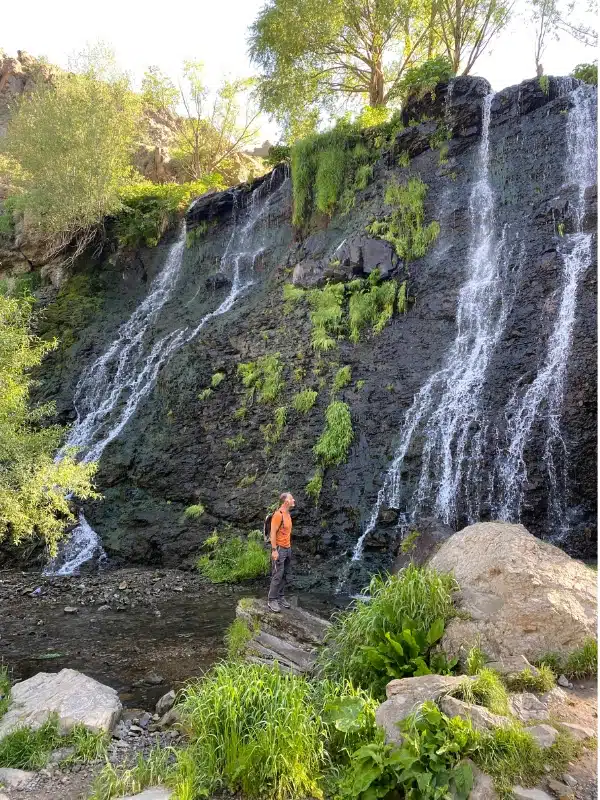 man in an orange t-shirt standing on a rock looking at a waterfall