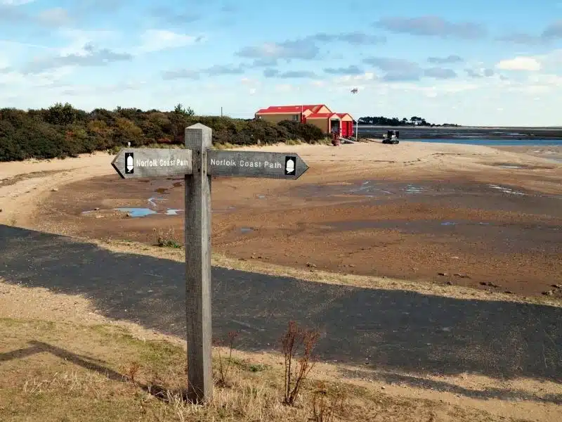 wooden 'Norfolk Coast Path' sign with a lifeboat house in the background