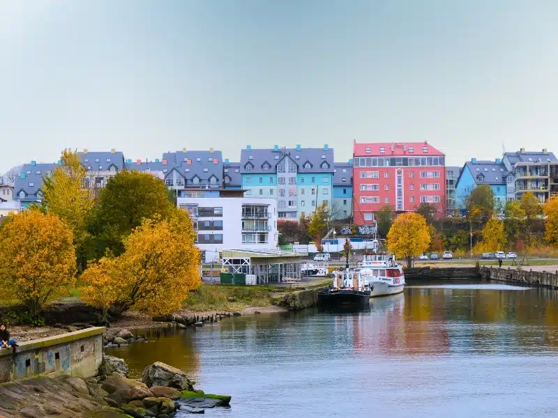 A calm waterway with boats docked along the shore, surrounded by vibrant autumn foliage and colorful buildings