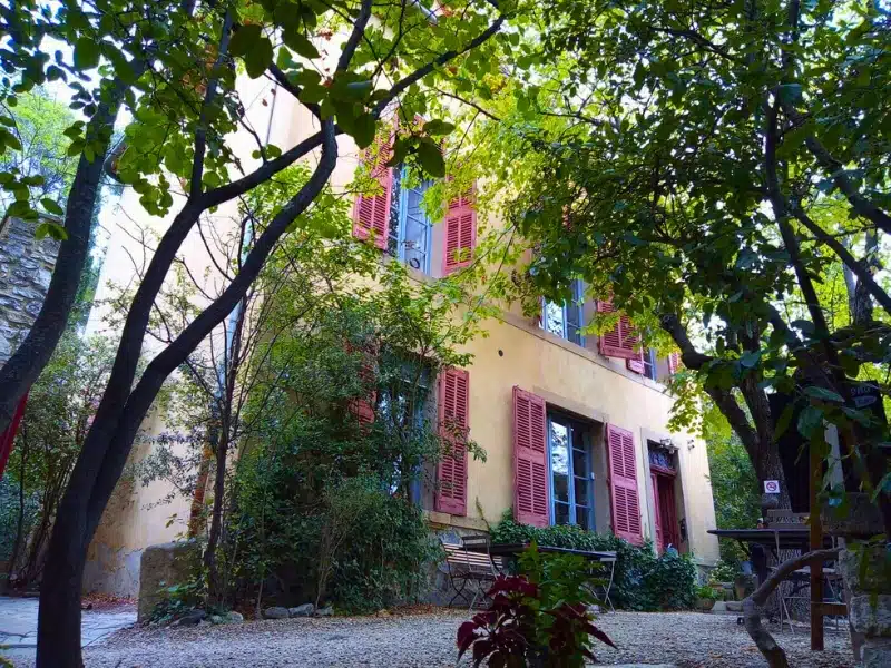 Town house in Aix-en-Provence painted a peachy colour with blue windows and red shutters, surrounded by trees.