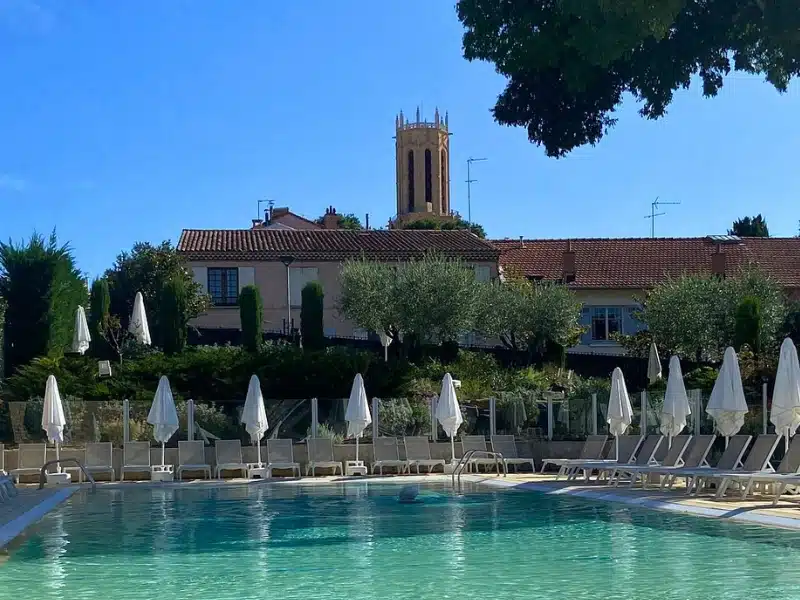 Swimming pool lined with sunbeds and umberellas and a red tiled Provencal building with tower in the background