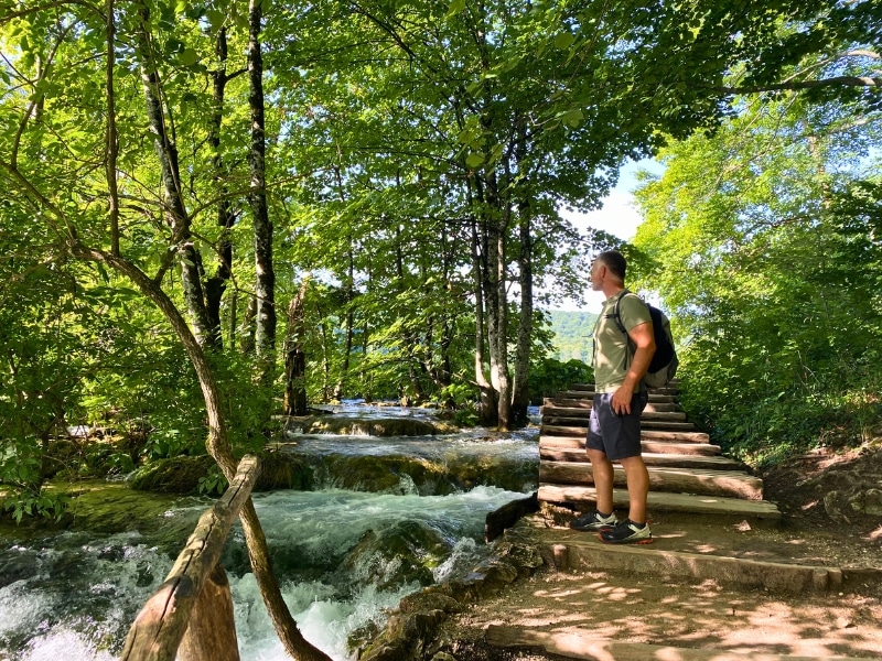 man with a rucksack standing on wooden stairs in nature looking at trees and rushing water by the path