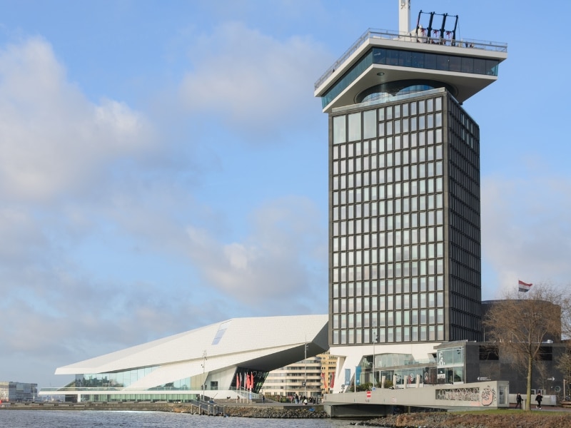 A'DAM Tower, a modern high-rise building with slanted glass panels, overlooking the Ij River in Amsterdam. In the foreground is a pier with docked boats and restaurants with outdoor seating.