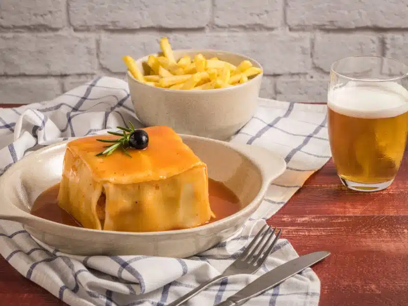 Traditional Portuguese snack food. Francesinha sandwich of bread, cheese, pork, ham, sausages, with tomato beer sauce and French fries. With beer and potatoes. On table.